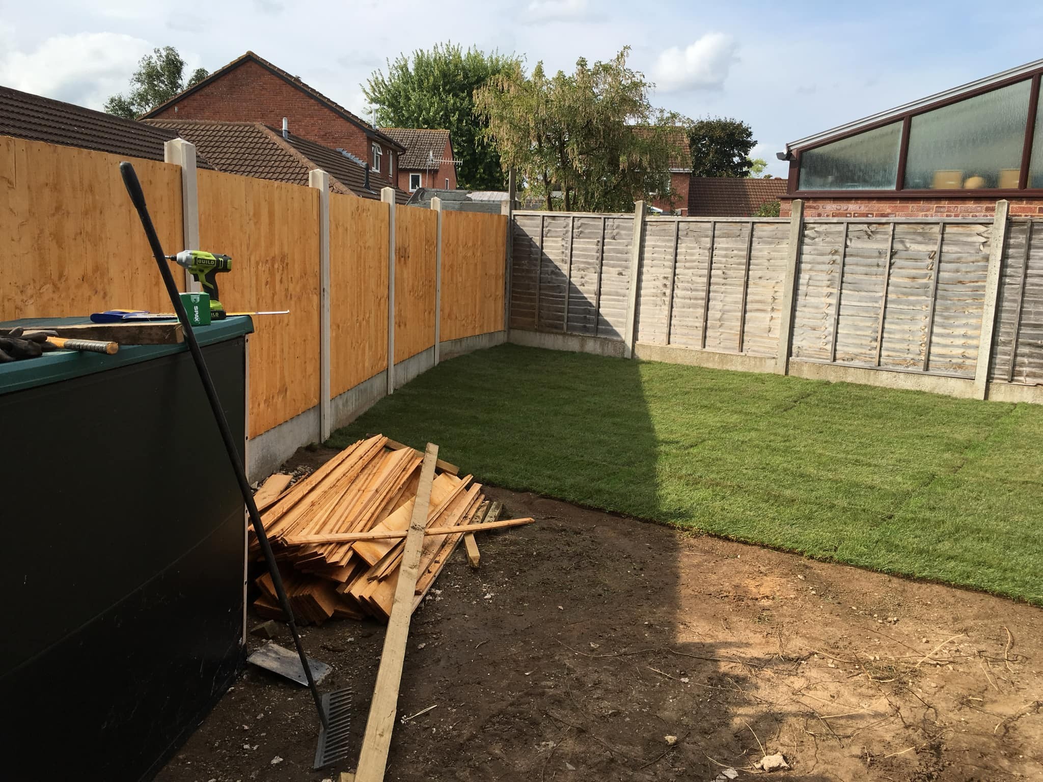 A backyard with new grass turf partially laid, wooden fencing installation on three sides, a pile of wooden planks, garden tools, and an oil tank on the left. The sky is clear and houses are visible in the background.