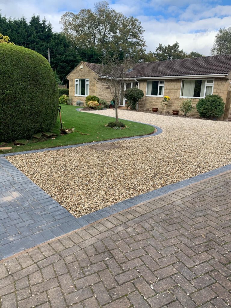 A single-story house with tan brick walls and a neatly landscaped front yard, featuring expert garden design, a paved driveway bordered by gravel, trimmed hedges, green grass, and small potted plants near the entrance.