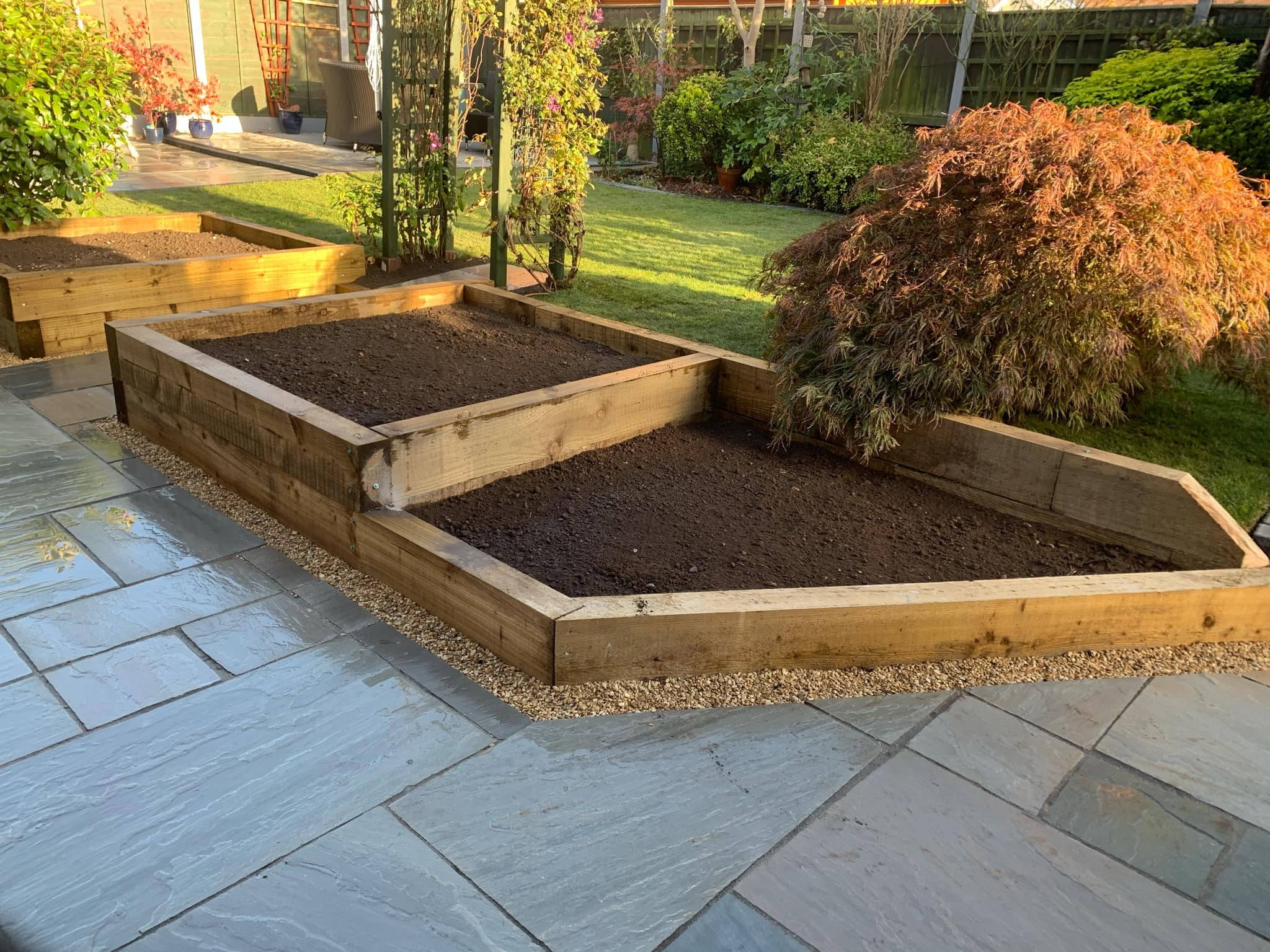 Raised wooden garden beds filled with soil sit beside a stone patio in a backyard landscaped by an expert landscaper; nearby are green shrubs, a maple tree, grass lawn, and additional plants along the fencing installation.