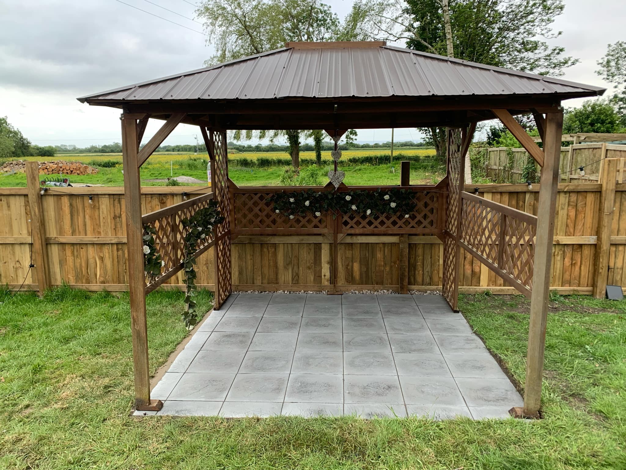 A wooden gazebo with a metal roof stands on a tiled patio, expertly crafted by a skilled landscaper, in a grassy yard surrounded by a wooden fence and trees, with open fields visible in the background.