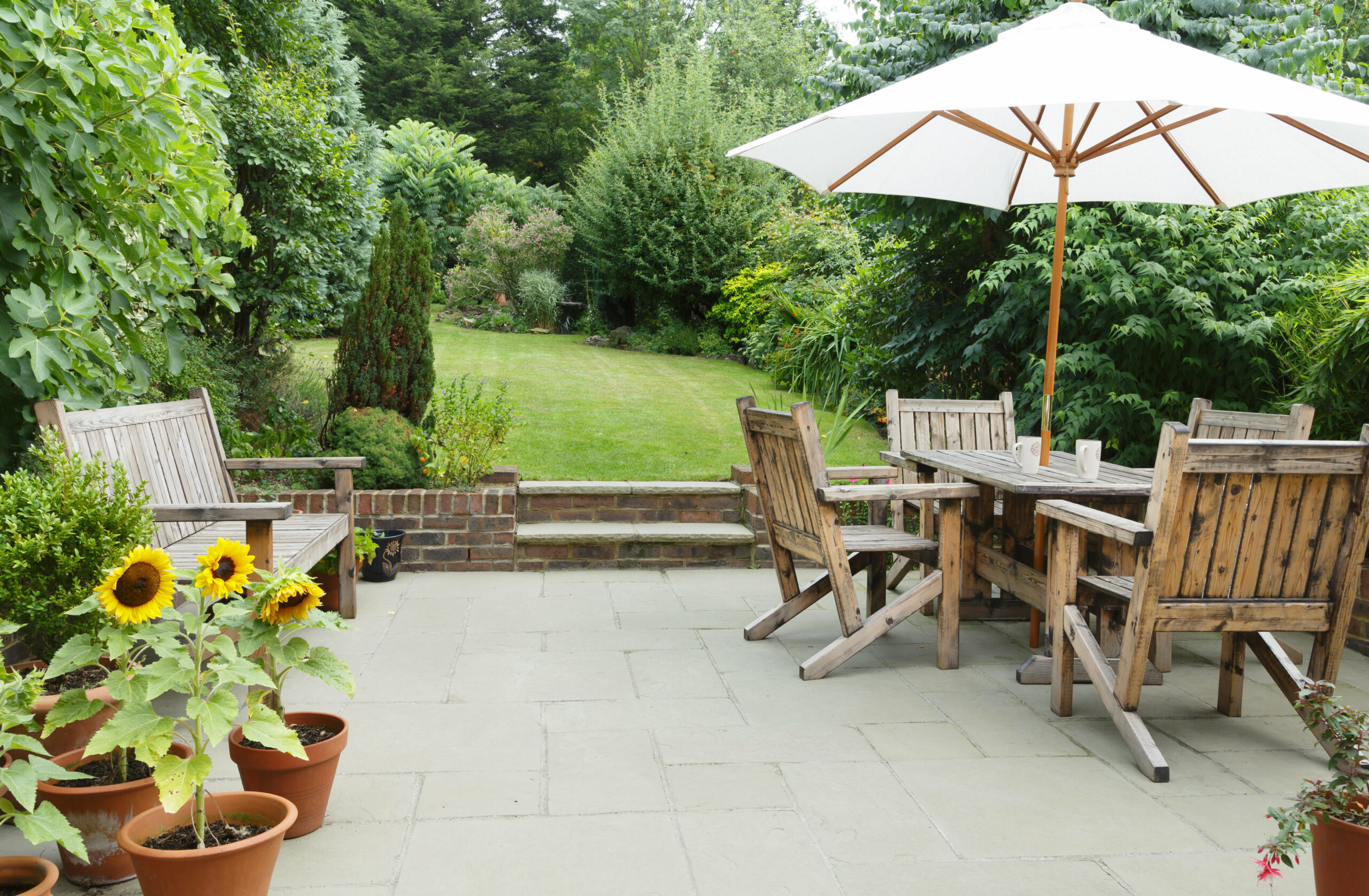 A patio with wooden benches and a dining table set under a large white umbrella, surrounded by potted plants and sunflowers, overlooks a lush green garden crafted by an expert landscaper, with trees and shrubs framing the serene space.