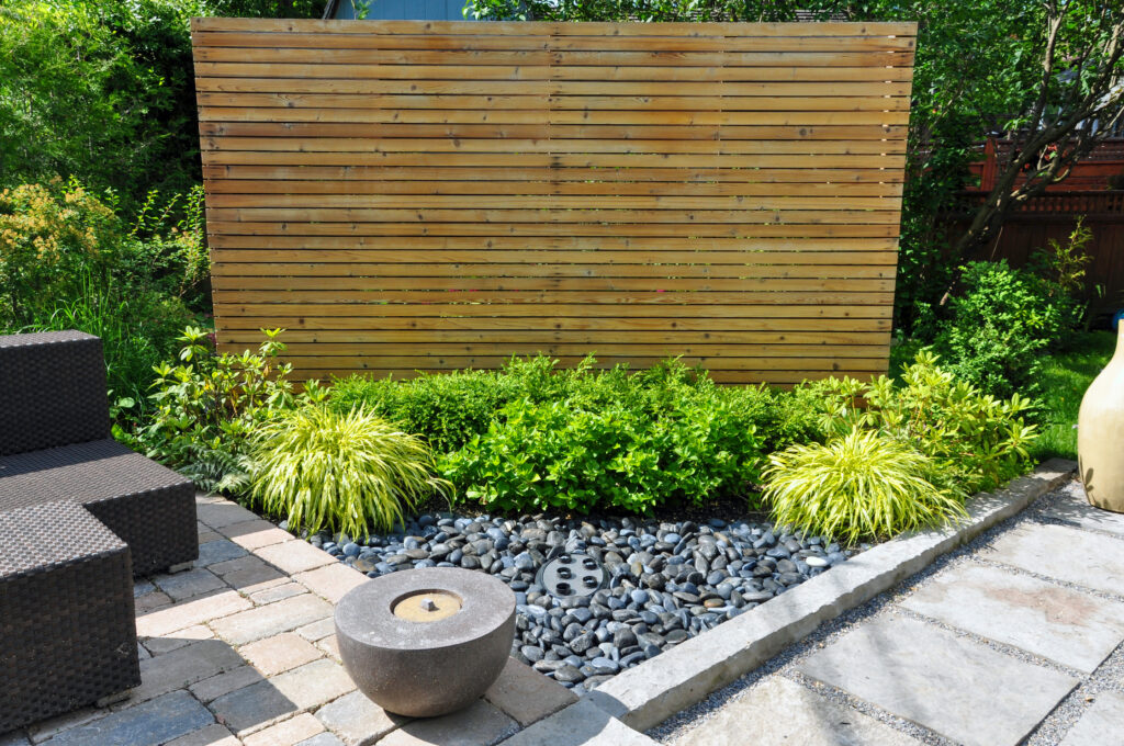 A modern backyard garden, crafted by an expert landscaper, features a wooden privacy screen, lush green shrubs, ornamental grasses, a small pebble bed, a round stone water fountain, and outdoor wicker furniture on a stone patio.