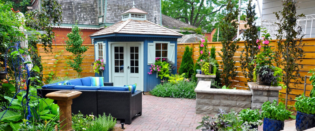 A vibrant backyard featuring professional patio laying on the brick patio, blue outdoor seating, lush plants, a birdbath, stone planter, and a small blue garden shed with white trim, all enclosed by expert fencing installation for added privacy.