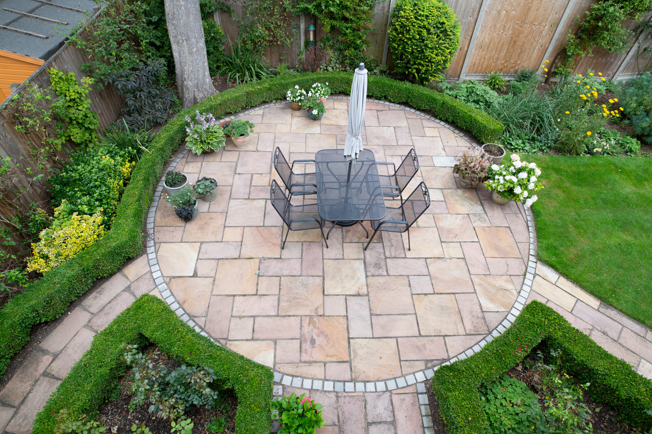 A circular stone patio with a black metal table, six chairs, and a closed umbrella is surrounded by neatly trimmed hedges, expert garden design, green lawns, and fencing installation in the backyard.