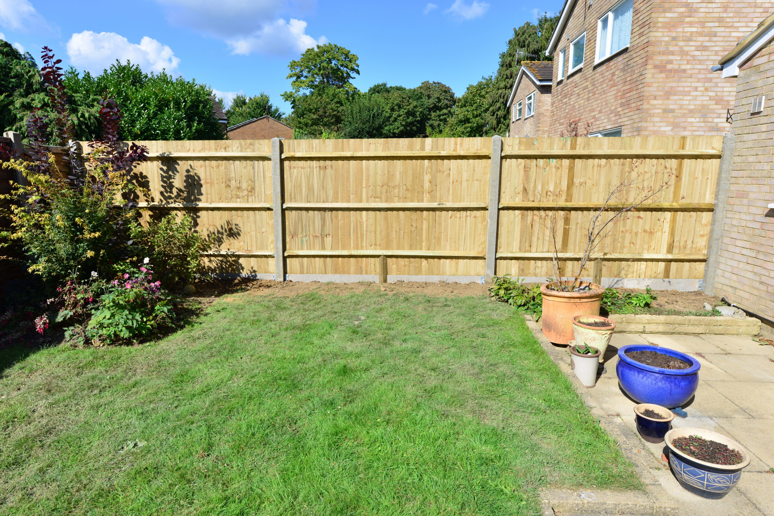 A backyard with a wooden fence from recent fencing installation, green lawn, and patio area with several potted plants. Shrubs and trees line the left side, a brick house is on the right, and clouds dot the blue sky.