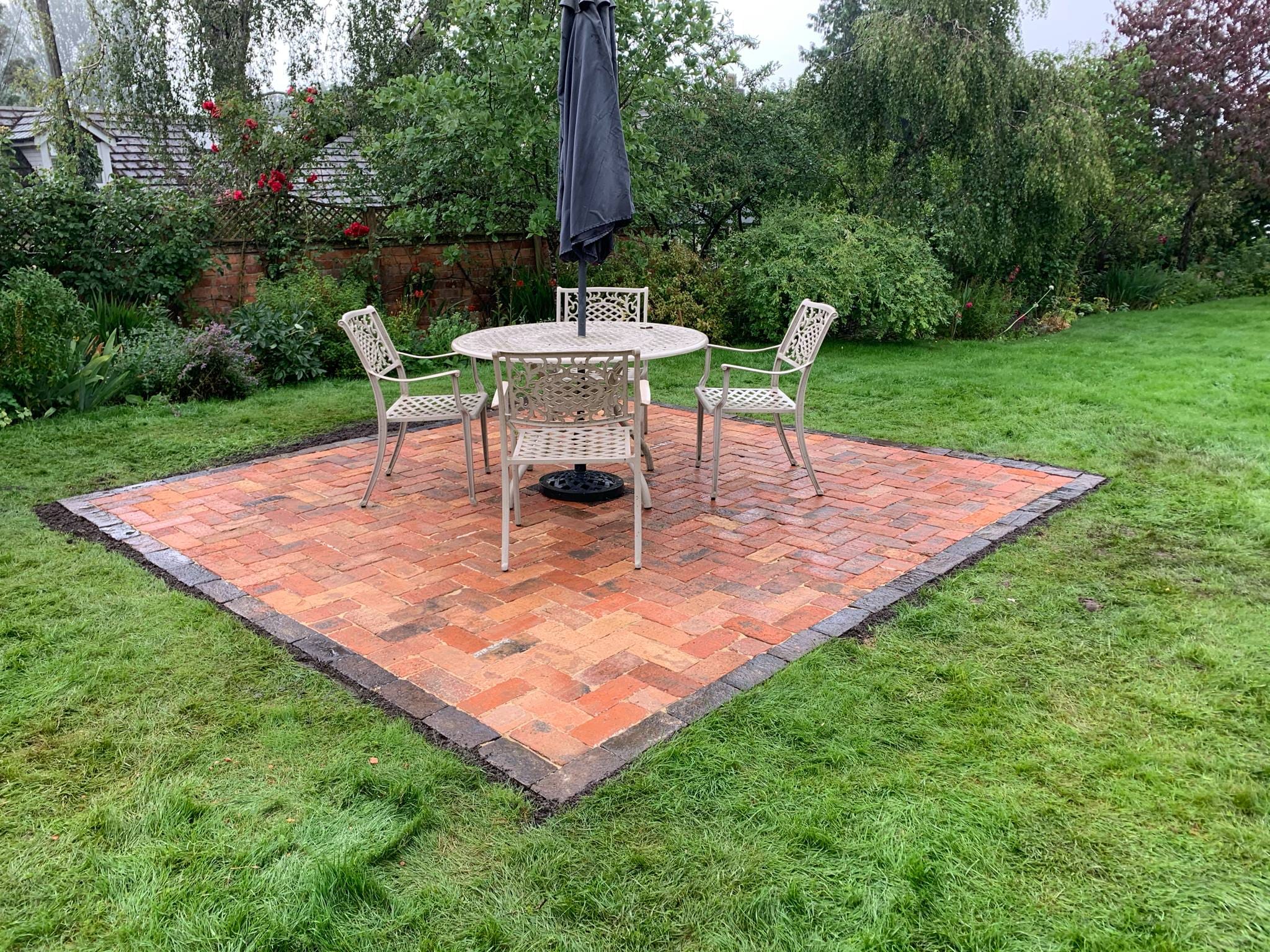 A small patio, crafted with expert patio laying, features a table, four chairs, and a closed umbrella atop red and brown bricks in a green backyard surrounded by trees and shrubs.